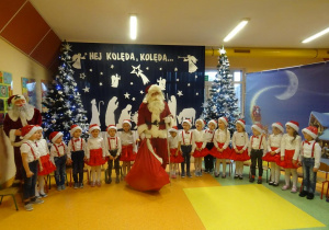 Mikołaj stoi pomiędzy dziećmi, w tle świąteczna dekoracja.